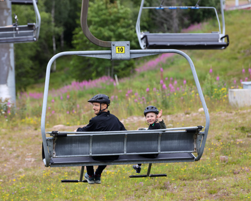 En vuxen och ett barn med cykelhjälmar sitter i en stollift på väg uppför en grön sluttning med blommande rallarrosor i bakgrunden.