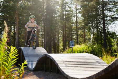 Barn med hjälm cyklar på en vågformad träbana i ett solbelyst skogsparti.