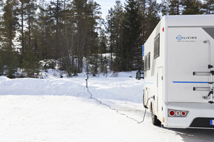 Autocamper parkeret i et snedækket landskab med strømkabel tilsluttet en elstander, omgivet af fyrreskov.