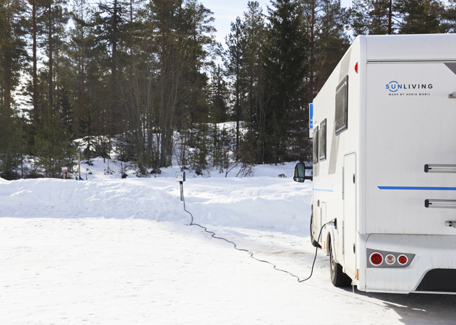 Autocamper parkeret i et snedækket landskab med strømkabel tilsluttet en elstander, omgivet af fyrreskov.