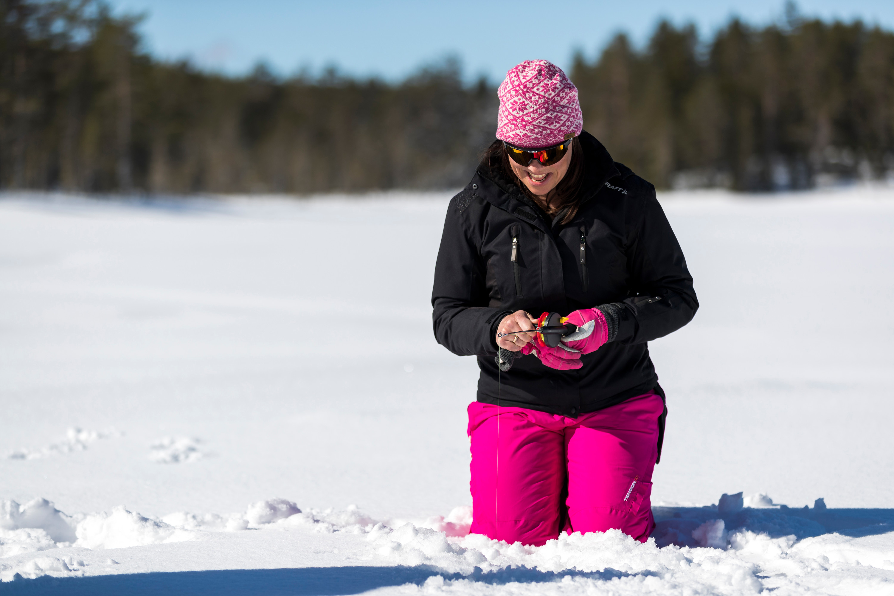 Kvinna klädd i svart jacka och rosa byxor som pimplar på en snötäckt sjö i solsken.