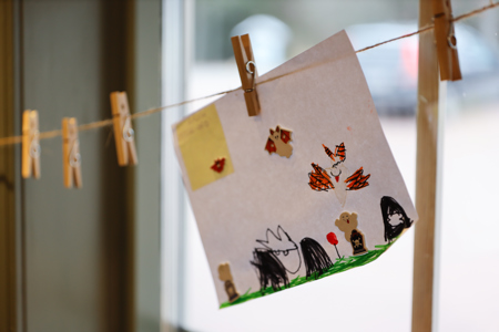 Ett barns teckning med färgglada figurer och klistermärken hänger i en klädnypa på ett snöre inomhus.