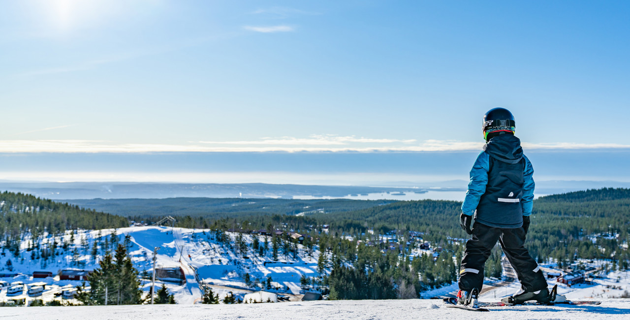 Child with helmet and skis stands on a snowy slope, looking out over the landscape in the winter sun.