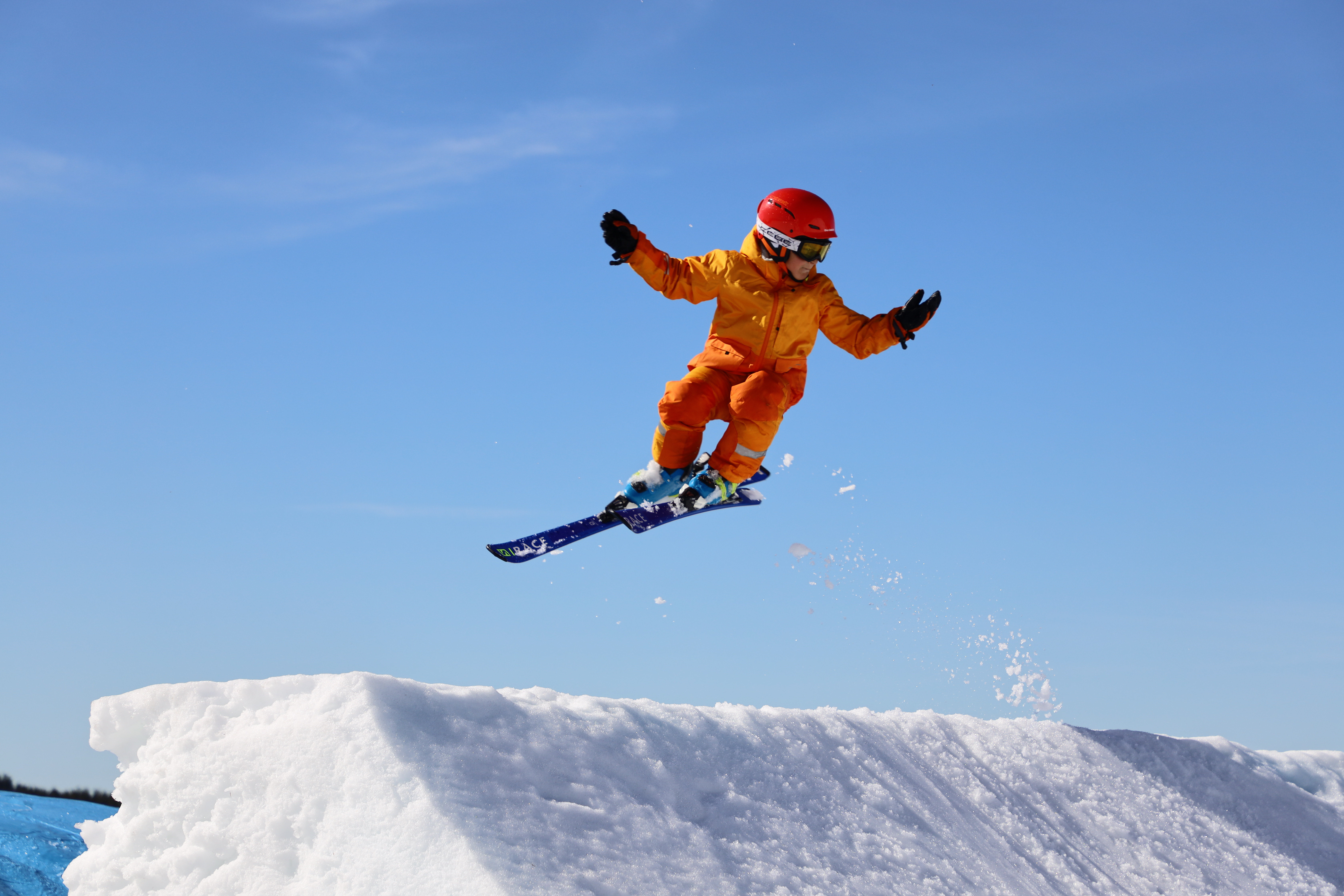 Ett barn i orange skidkläder och röd hjälm gör ett hopp med skidor i en snowpark mot klarblå himmel.