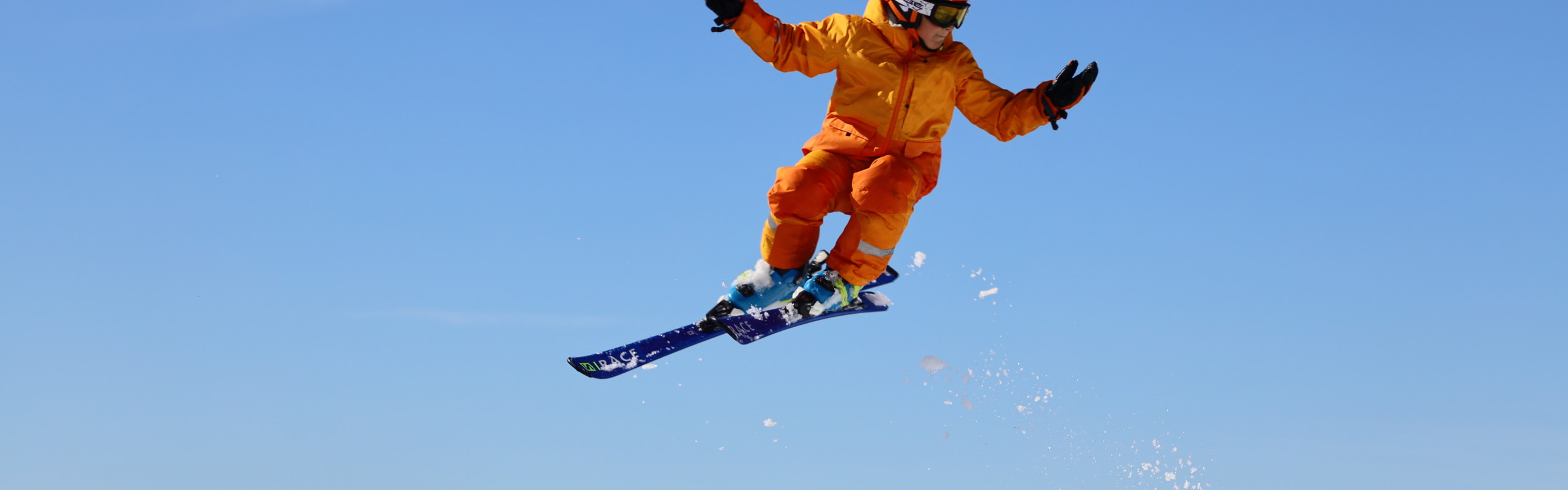 Ett barn i orange skidkläder och röd hjälm gör ett hopp med skidor i en snowpark mot klarblå himmel.