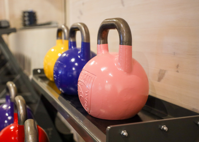 Flera färgglada kettlebells, bland annat rosa, blå och gula, står uppradade på ett hyllställ i ett gym.