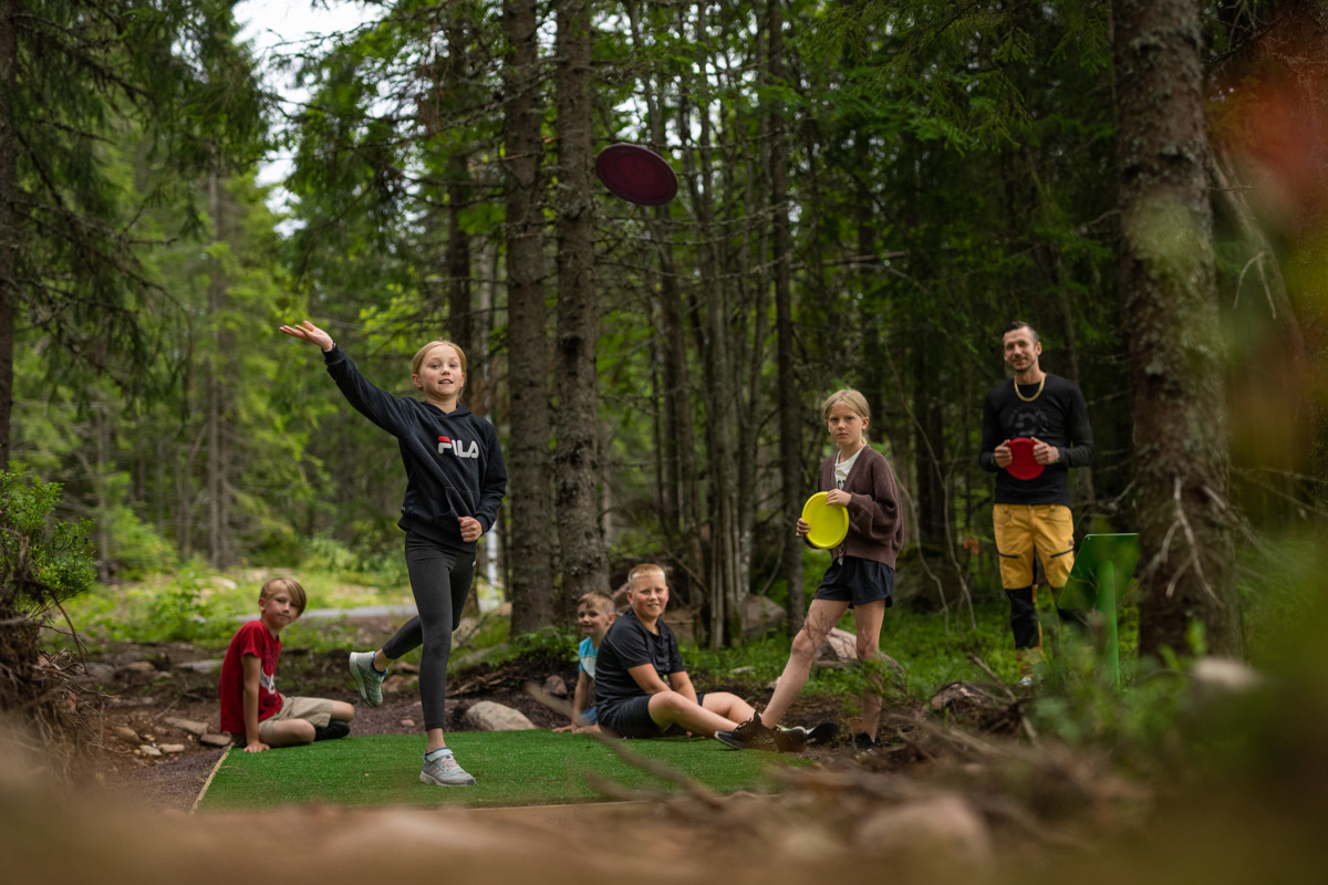 Barn och vuxna spelar discgolf i en skogsglänta, mitt i ett kastögonblick.