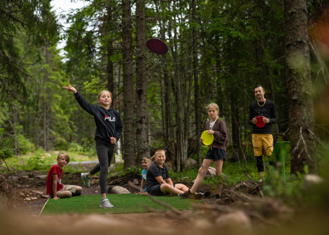 Barn och vuxna spelar discgolf i en skogsglänta, mitt i ett kastögonblick.