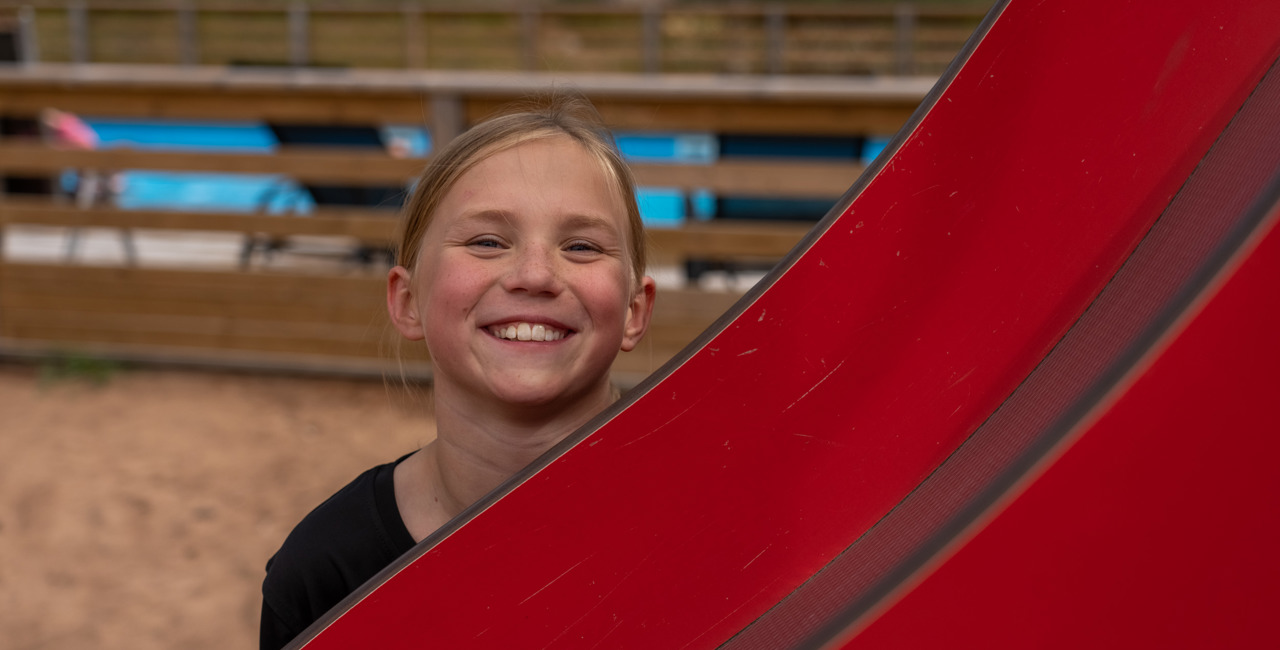 Ett leende barn tittar fram bakom en röd vattenrutschkana vid ett poolområde med trästaket i bakgrunden.
