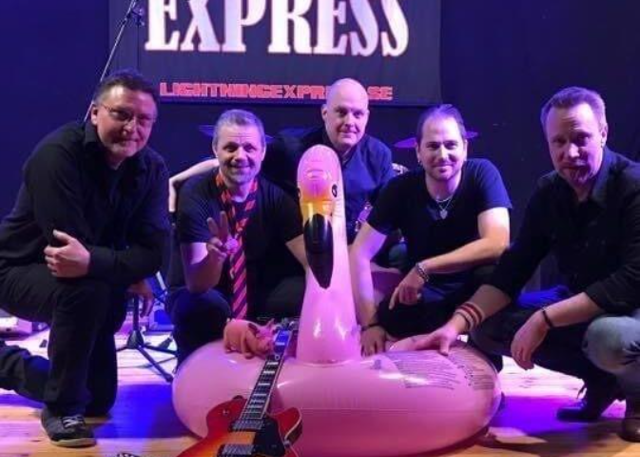 Fem musiker poserar på en scen framför en skylt med texten ”Lightning Express”, tillsammans med en uppblåsbar rosa flamingo och en elgitarr på golvet.
