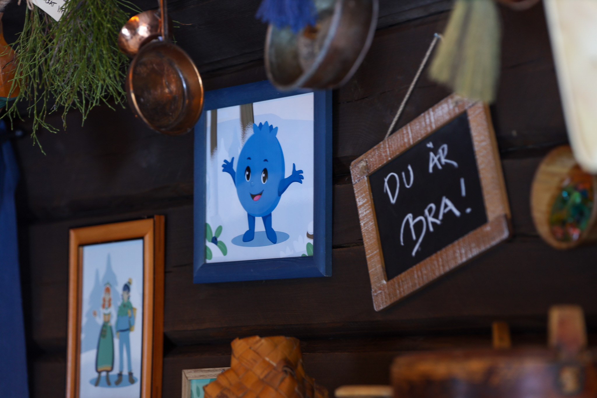 Vägg med tavlor och dekorationer, där en illustration av Blåbäret Bärra och en liten griffeltavla med texten ”Du är bra!” hänger bland koppar och kvistar.
