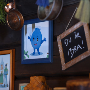 Vägg med tavlor och dekorationer, där en illustration av Blåbäret Bärra och en liten griffeltavla med texten ”Du är bra!” hänger bland koppar och kvistar.