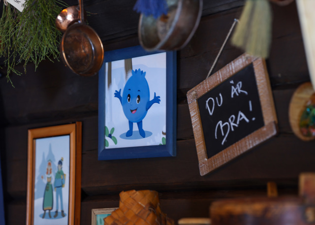 Vägg med tavlor och dekorationer, där en illustration av Blåbäret Bärra och en liten griffeltavla med texten ”Du är bra!” hänger bland koppar och kvistar.