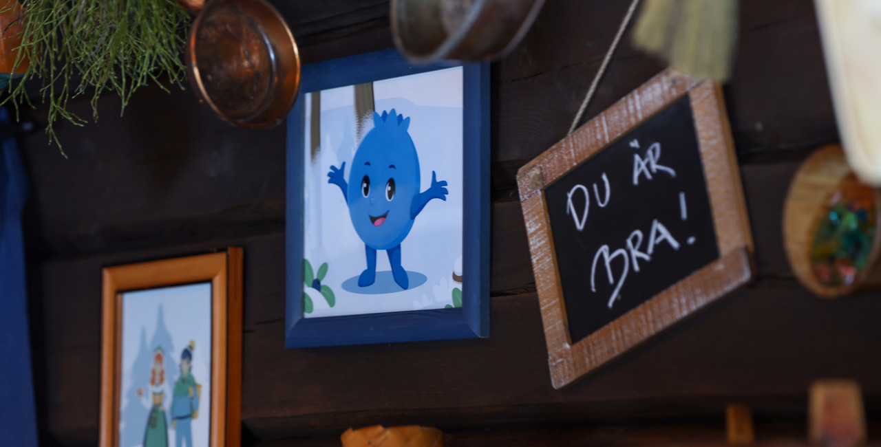 Wall decorated with framed pictures, including an illustration of Blåbäret Bärra and a small chalkboard reading “You are great!”, surrounded by copper items and hanging greenery.