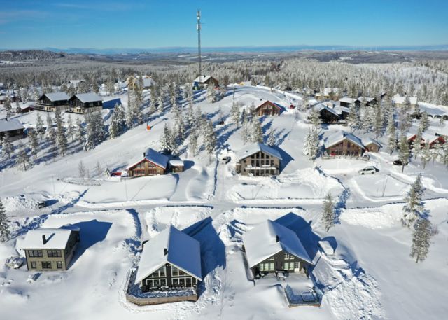 Luftfoto af moderne fjeldhytter i snedækket landskab med mast i baggrunden.