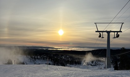 Snötäckt skidbacke med liftstolpar, snökanoner och låg sol över landskapet.