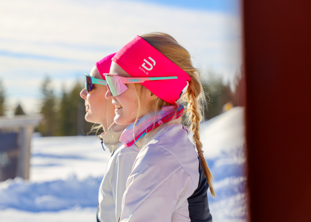 To piger med farverige pandebånd og solbriller nyder solen på en vinterdag i skiomgivelser.