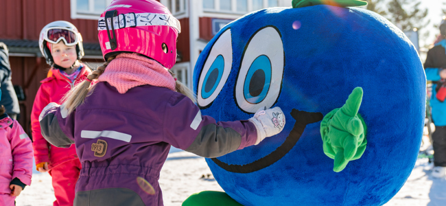 Maskoten Bärra hälsar på barn i skidkläder utanför en röd byggnad i vintrig miljö.