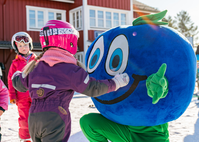 Maskoten Bärra hälsar på barn i skidkläder utanför en röd byggnad i vintrig miljö.