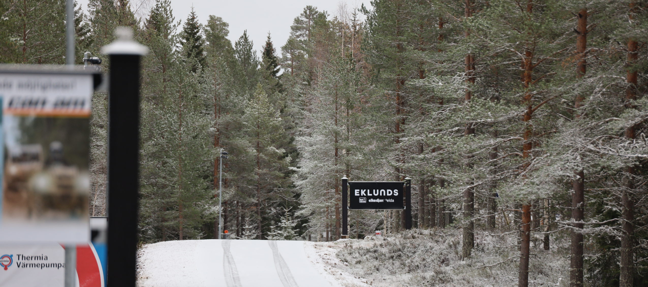 Snötäckt väg som leder in i en tallskog med en skylt för Eklunds på högra sidan.