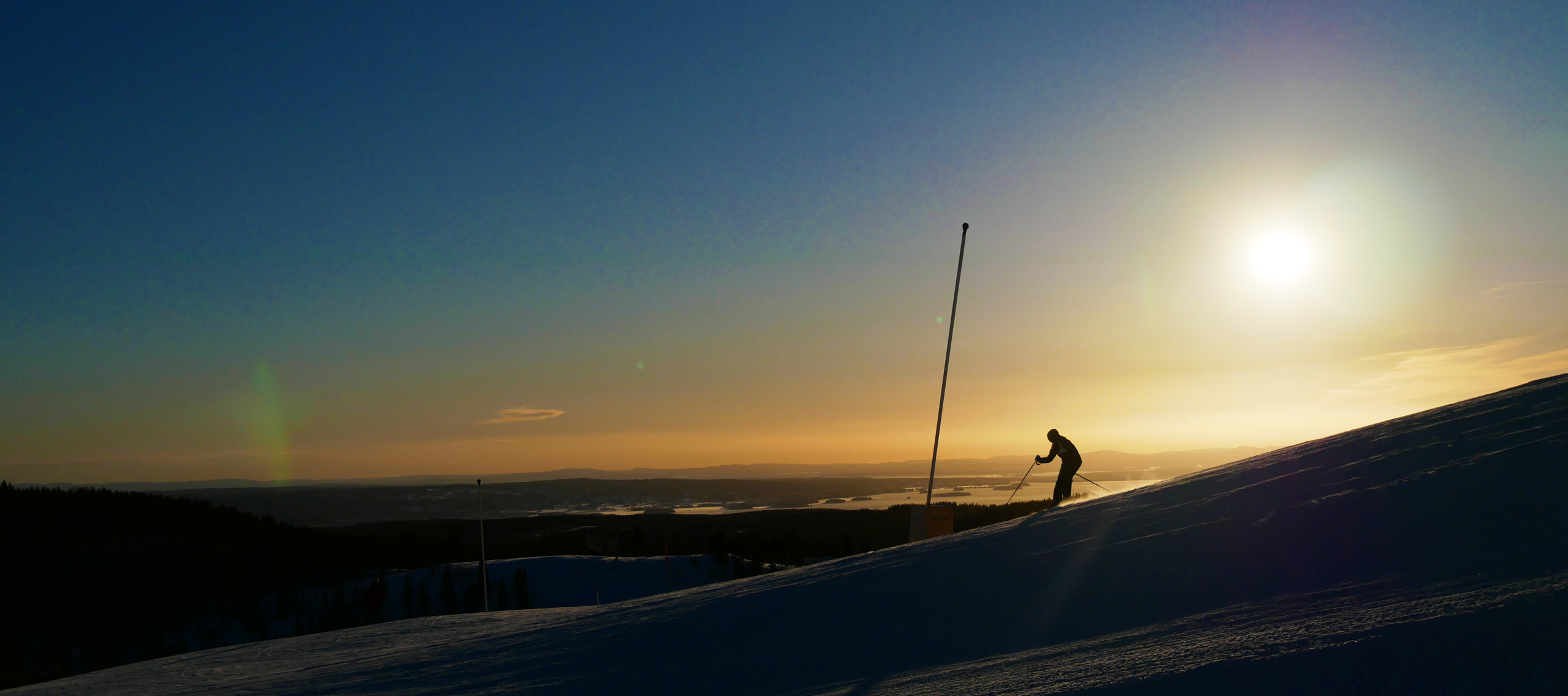 Skidåkare tar sig uppför en pist i solnedgång med utsikt över ett snötäckt landskap.