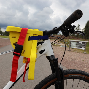Närbild på ett färgglatt barncykelsäte i gult, rött och blått, monterat framtill på en vit mountainbike, med grusväg och skog i bakgrunden.