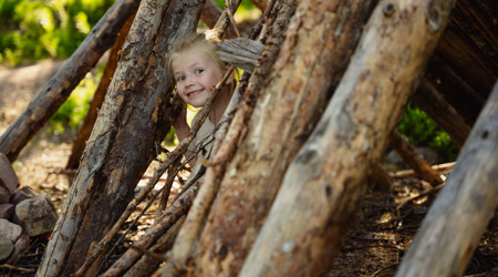 Litet barn kikar leende ut genom en koja byggd av stora trästockar i skogen.