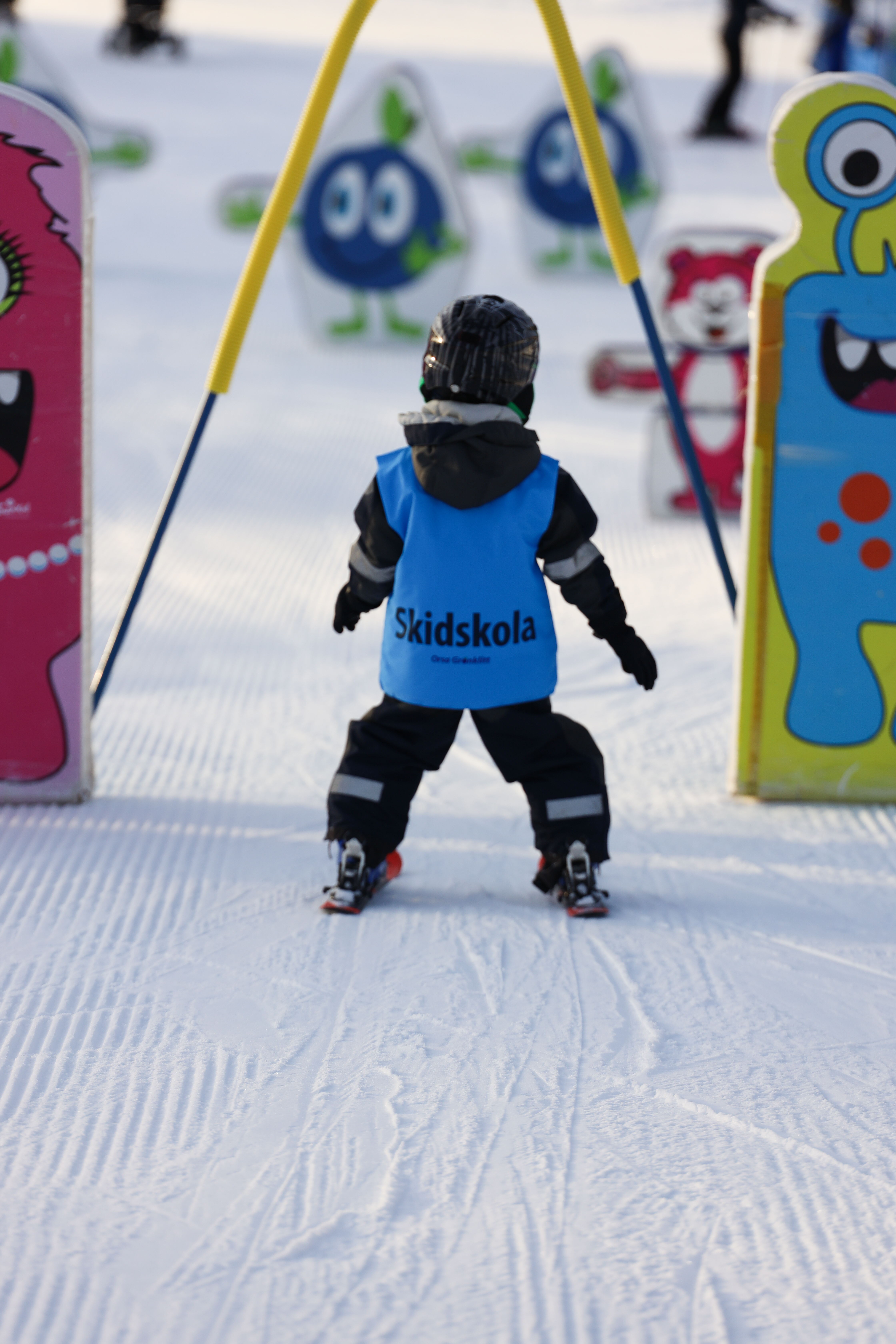 Litet barn med blå väst märkt ”Skidskola” åker mellan färgglada figurer i barnområdet.
