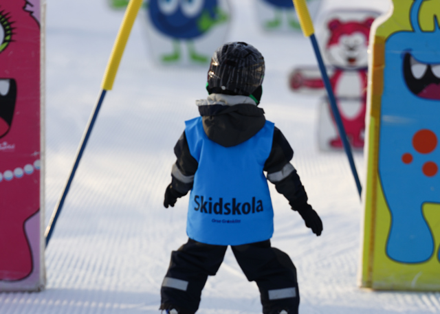 Small child wearing a blue vest labeled “Ski school” skiing between colorful figures in the kids’ area.