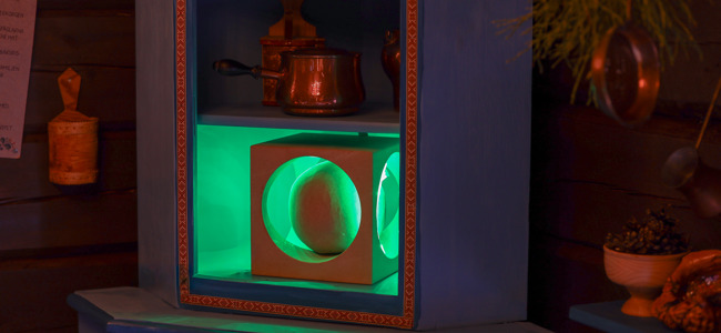Hylla med ett belyst grönt fack där ett runt föremål står i en kvadratisk ram, omgiven av köksföremål i en träinteriör.