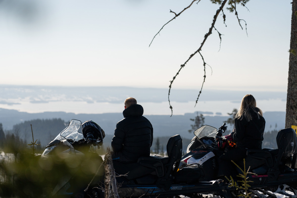 Två personer sitter vid sina snöskotrar och njuter av utsikten över landskapet.