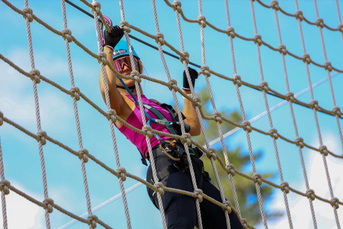 En kvinde med sikkerhedssele klatrer i et rebnet i en højderute og smiler til kameraet.
