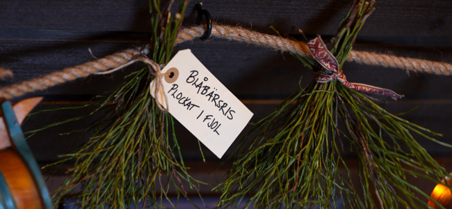 Bundna kvistar hängande i ett snöre med en etikett som texten ”Blåbäret plockat i fjol”, i en träinteriör.