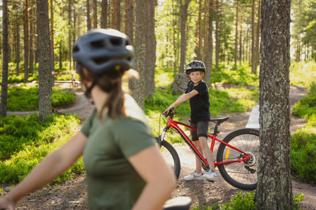 Boy with a red bike and helmet stands in the forest, with an adult visible in the foreground.
