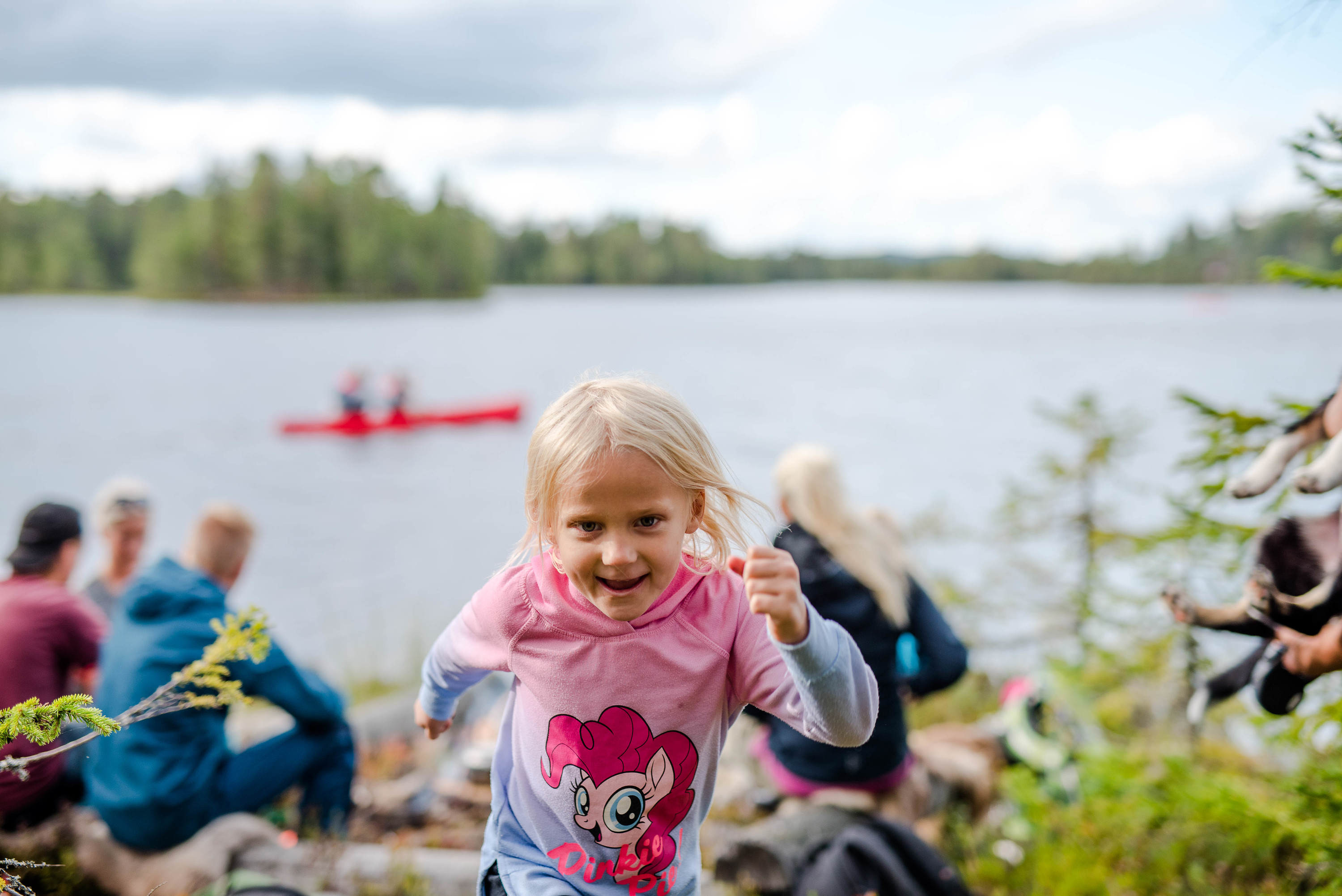 Girl in a pink sweater running toward the camera by a lake, with canoe paddlers and other children in the background.