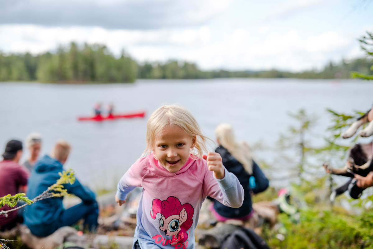 Girl in a pink sweater running toward the camera by a lake, with canoe paddlers and other children in the background.