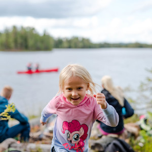 Girl in a pink sweater running toward the camera by a lake, with canoe paddlers and other children in the background.