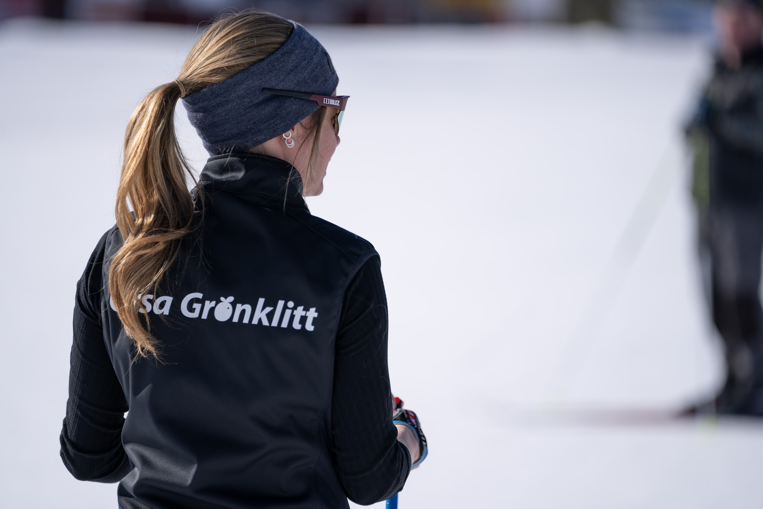 Person med hästsvans och svart väst med Orsa Grönklitt-logga står i snön med ryggen mot kameran.