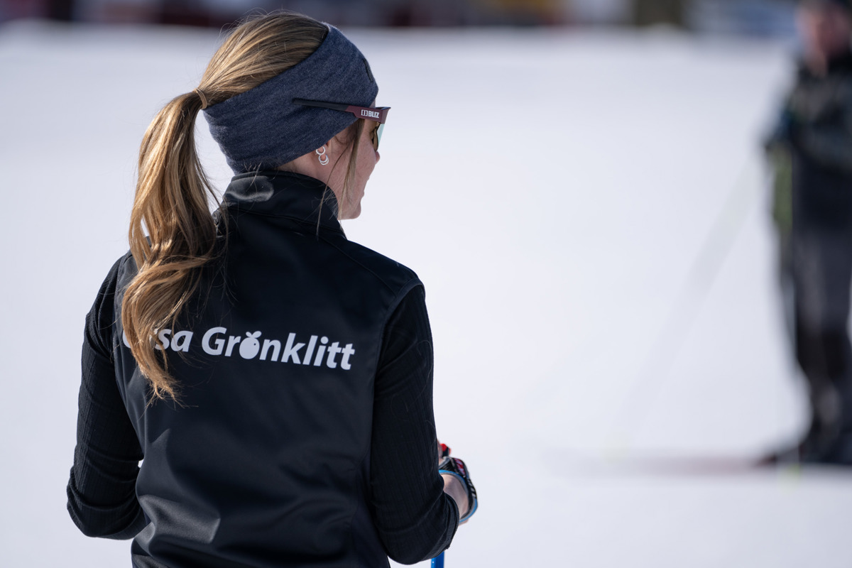 Person med hästsvans och svart väst med Orsa Grönklitt-logga står i snön med ryggen mot kameran.