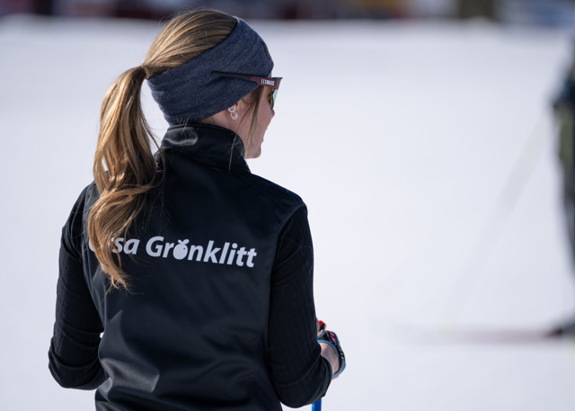 Person with a ponytail and black vest featuring the Orsa Grönklitt logo standing in the snow, facing away from the camera.