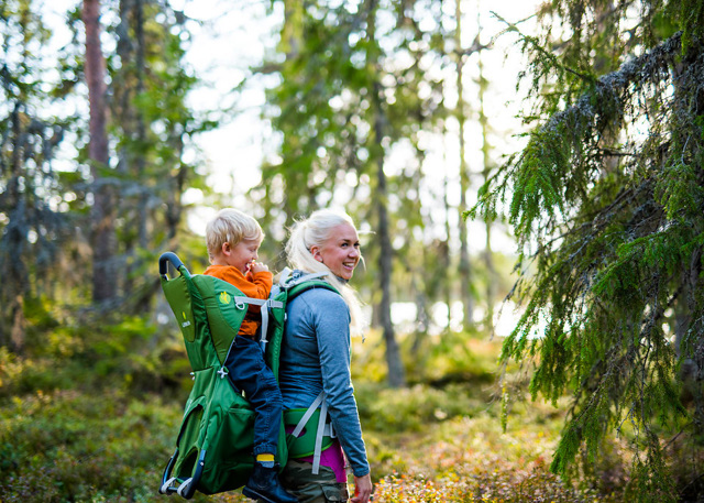 Kvinna vandrar i skogen med ett barn i bärstol på ryggen.
