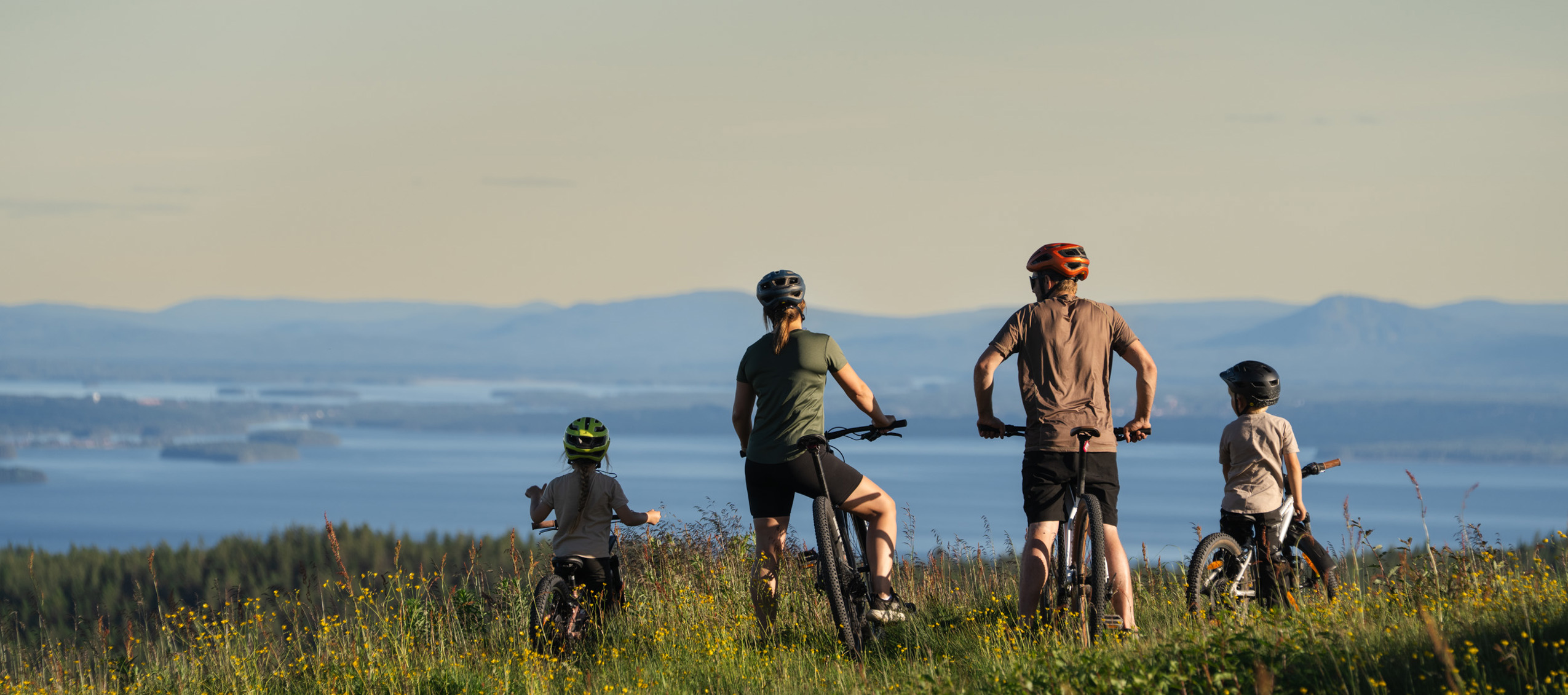 En familj med två vuxna och två barn står med sina cyklar i en blommande sommaräng och blickar ut över en vidsträckt utsikt med sjöar och blå berg i horisonten.