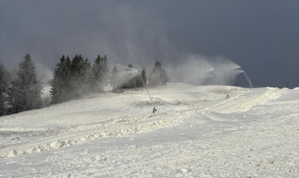 Snökanoner som producerar snö över ett vitt vinterlandskap med skog i bakgrunden och mörka moln på himlen.