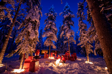 “Snow-covered winter forest at dusk with people dressed as Santa Claus. They sit by a campfire surrounded by large wrapped gifts and illuminated pine trees.