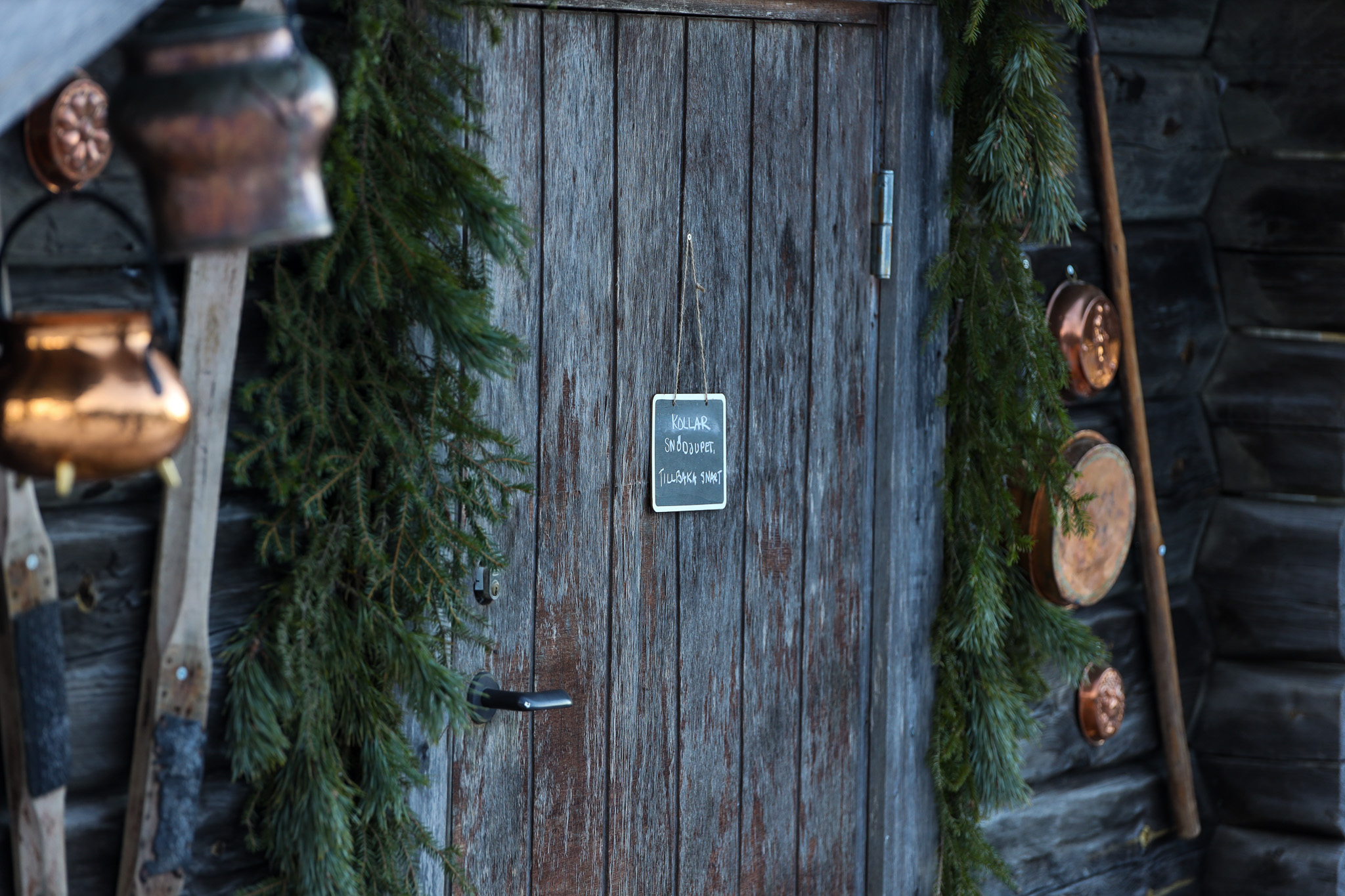 Trädörr dekorerad med granris och en liten griffeltavla med texten ”Kollar odjuret, tillbaka snart”, omgiven av kopparkärl och träredskap.