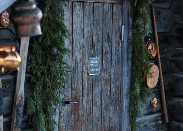 Trädörr dekorerad med granris och en liten griffeltavla med texten ”Kollar odjuret, tillbaka snart”, omgiven av kopparkärl och träredskap.